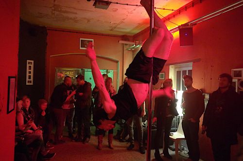 Foto, wie ich kopfüber mit den Beinen an der Pole klemme. Ringsum stehen und sitzen Leute. An den Wänden sind gerahmte Fotografien ausgestellt. Der Raum ist schummrig, rot-orange beleuchtet.