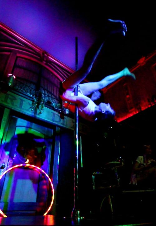 Foto von mir an der Pole. Rücken nach unten, Pole ist an Hüfte, zwischen den Beinen und klemmt in linker Achsel. Beine sind schräg nach vorn gestreckt. Die Füße sind angewinkelt. Der LED-Hoop lehnt hinten an der Wand. 